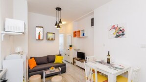 Un saló modern amb un sofà, otomana, taula de dinar i cadirees. Hi ha una televisió, una ampolla de vi i un quadre a la paret.