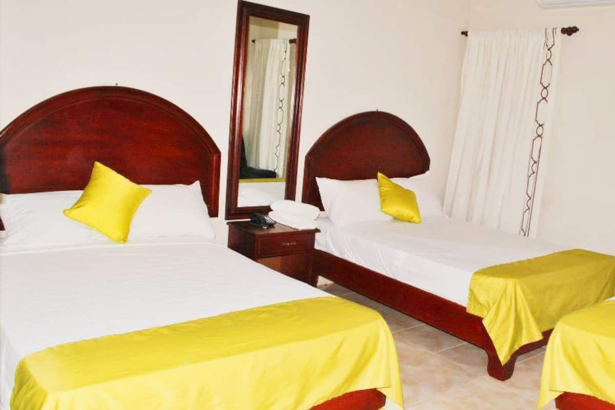 A hotel room with three single beds, each with a wooden headboard, white bedding, and yellow bedspreads. There is a nightstand with a telephone and a mirror on one side.