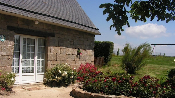 A stone building with a white door and windows, surrounded by a garden with various flowers and a well-maintained lawn. - Charming house in a quiet (Guengat)
