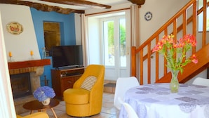 A cozy living room with a fireplace, a television, a yellow armchair, a dining table with a white tablecloth, and a vase of flowers. - Charming house in a quiet (Guengat)