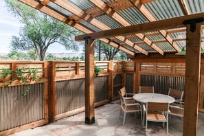 Un espacio de patio cubierto con una pérgola de madera, una mesa redonda con sillas, y vistas a árboles y montañas al fondo.