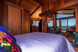 Ein geräumiges Schlafzimmer mit einem großen Bett, Holzbalkendecke und Meerblick durch große Fenster.