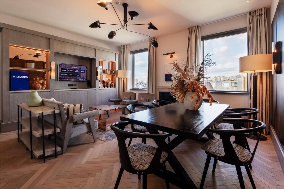 A modern hotel room with a dining area, a TV mounted on the wall, and a large window offering a city view.