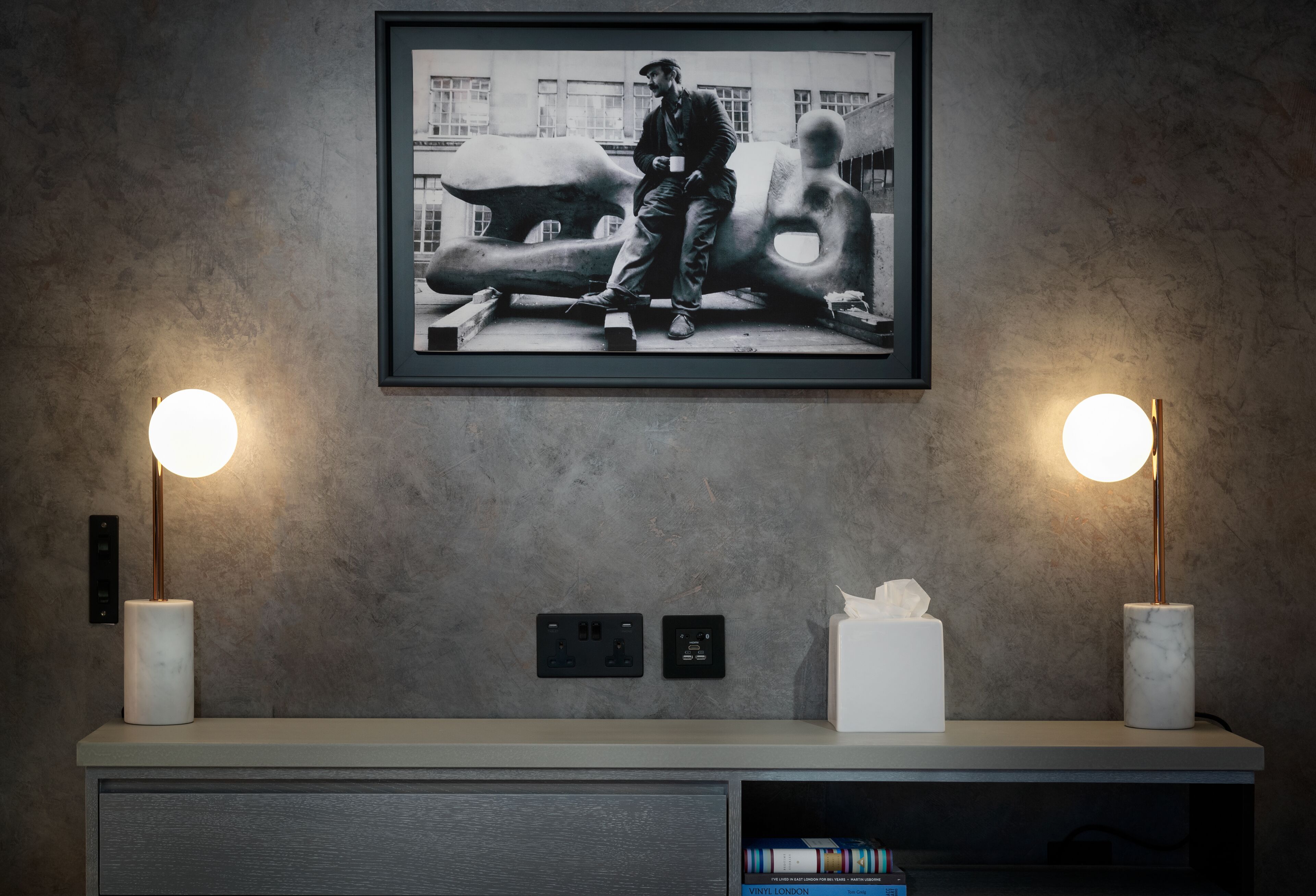 A modern room with a large framed photograph on the wall, two wall-mounted lamps, a white tissue box, and a built-in bookshelf with books.