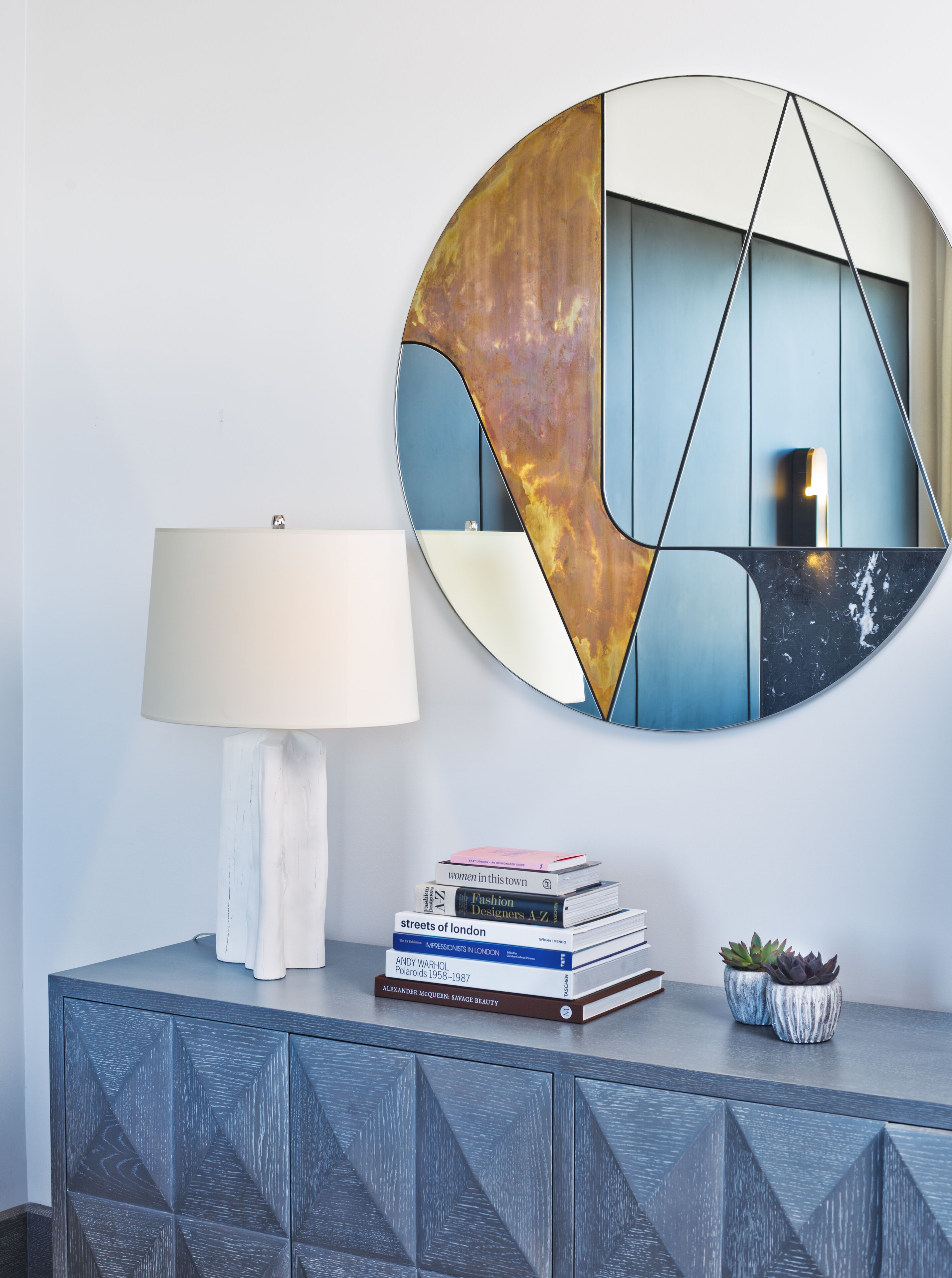 A modern room with a geometric-patterned cabinet, a stack of books, a lamp, and a decorative wall piece.
