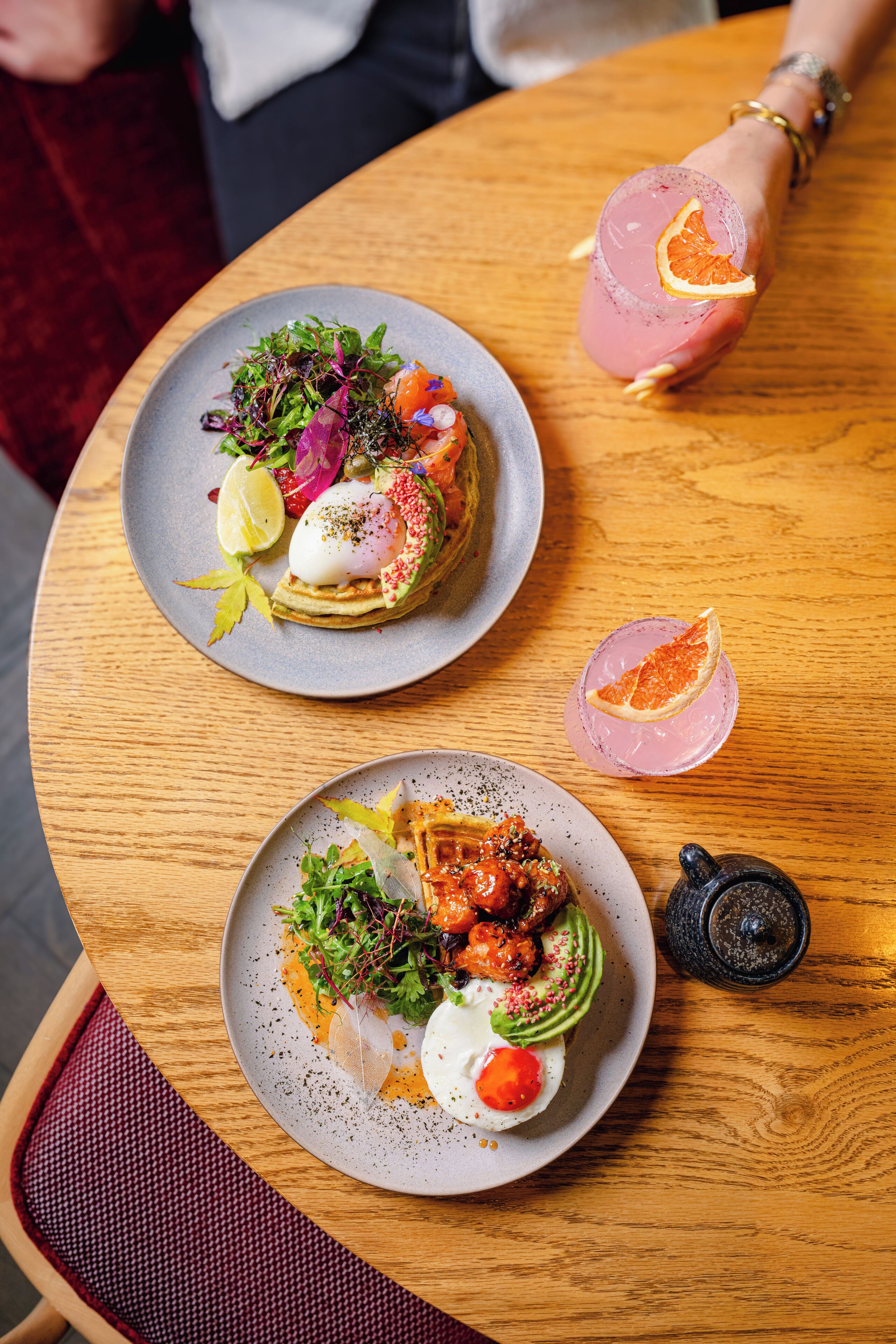 Two plates of food on a wooden table, one with a salad and the other with a dish that includes a poached egg and avocado.