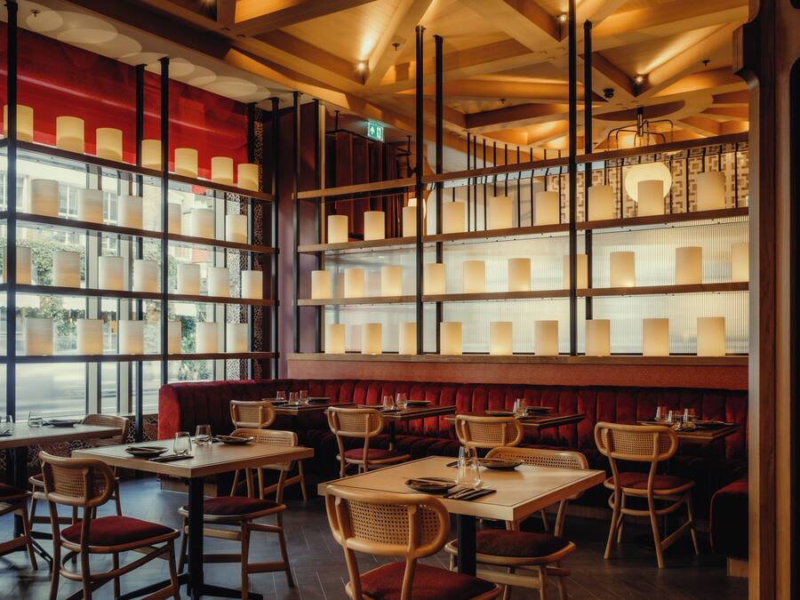 A restaurant interior with multiple tables and booths, wooden chairs, and a red cushioned bench area.