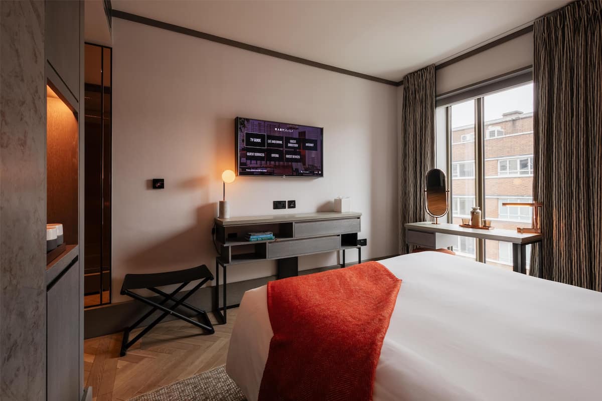 A hotel room with a bed, a desk with a flat-screen TV, a chair, and a window with curtains.