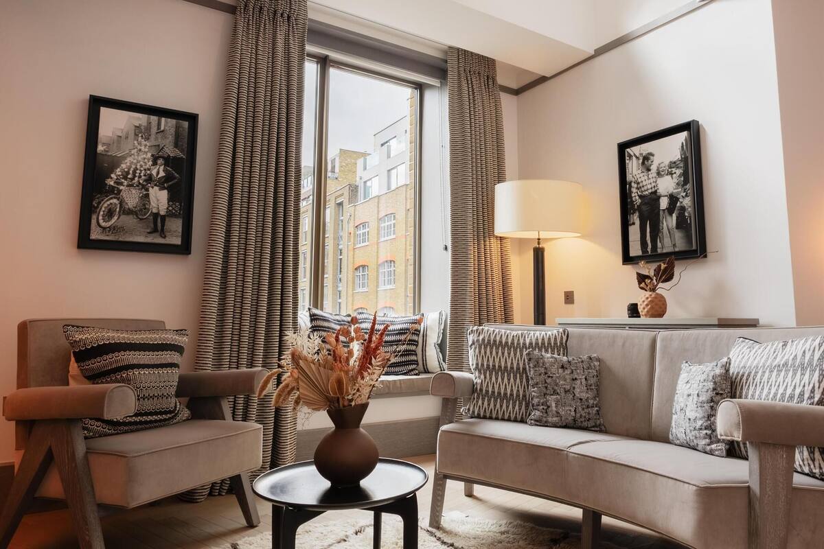 A modern living room with a high ceiling, a geometric pendant light, a large window with curtains, and two sofas with patterned cushions.