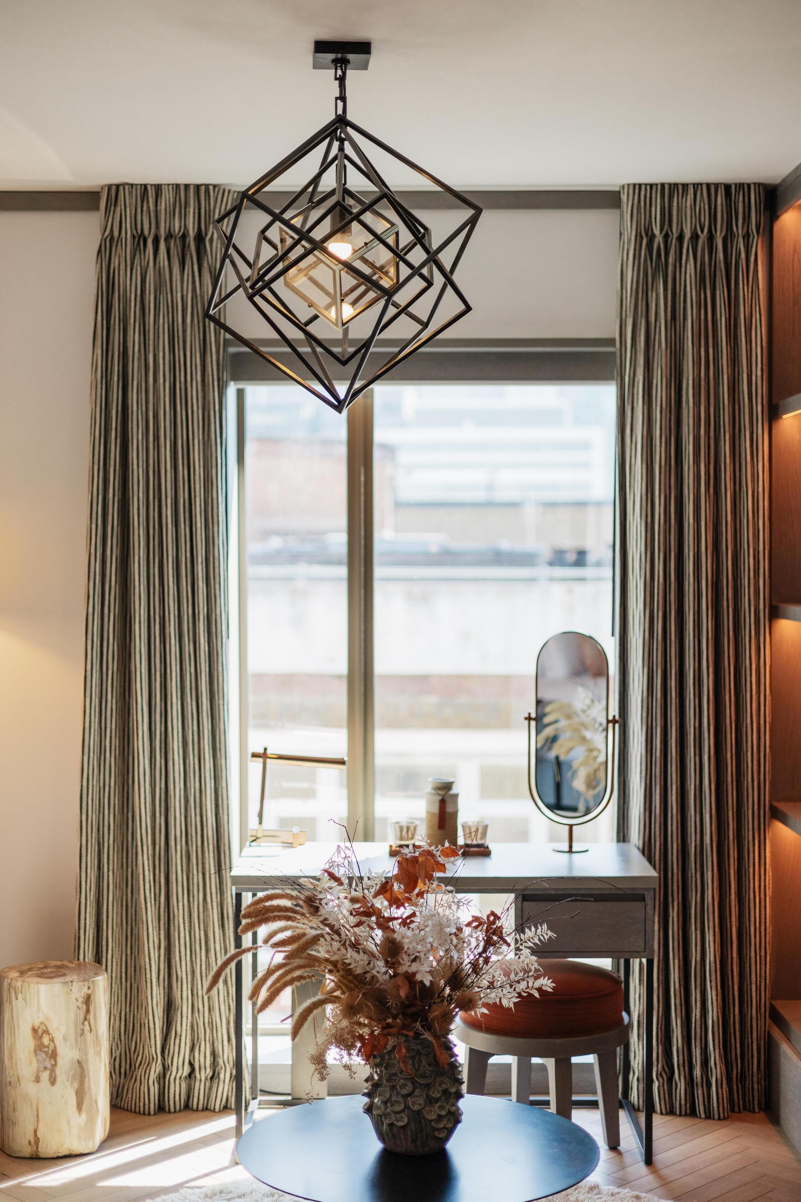 A modern room with a geometric pendant light, a round table with a vase of dried flowers, a mirror, and striped curtains.