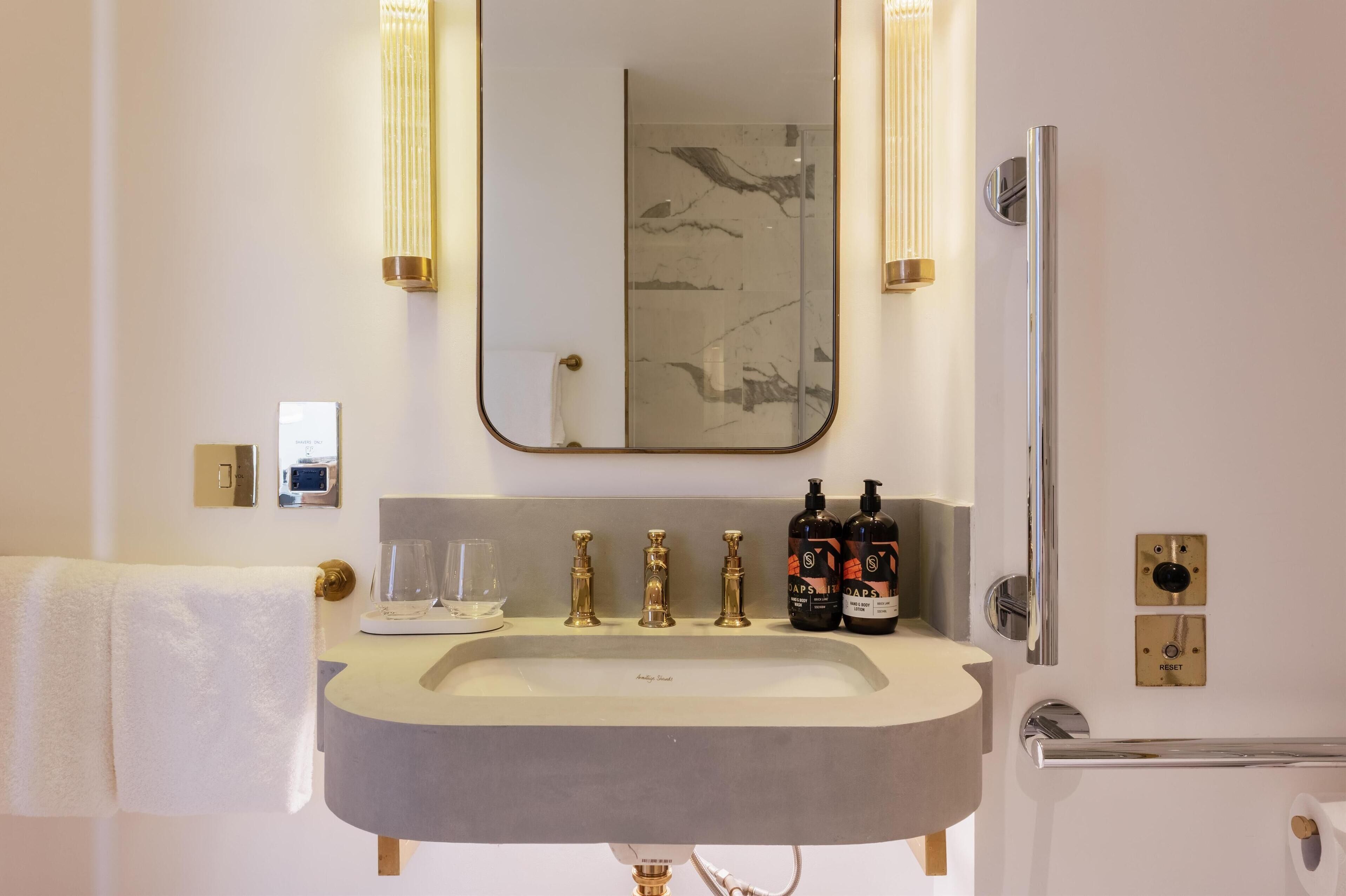 A bathroom with a large mirror, a sink with three faucets, two bottles of soap, and a towel hanging on a rack.