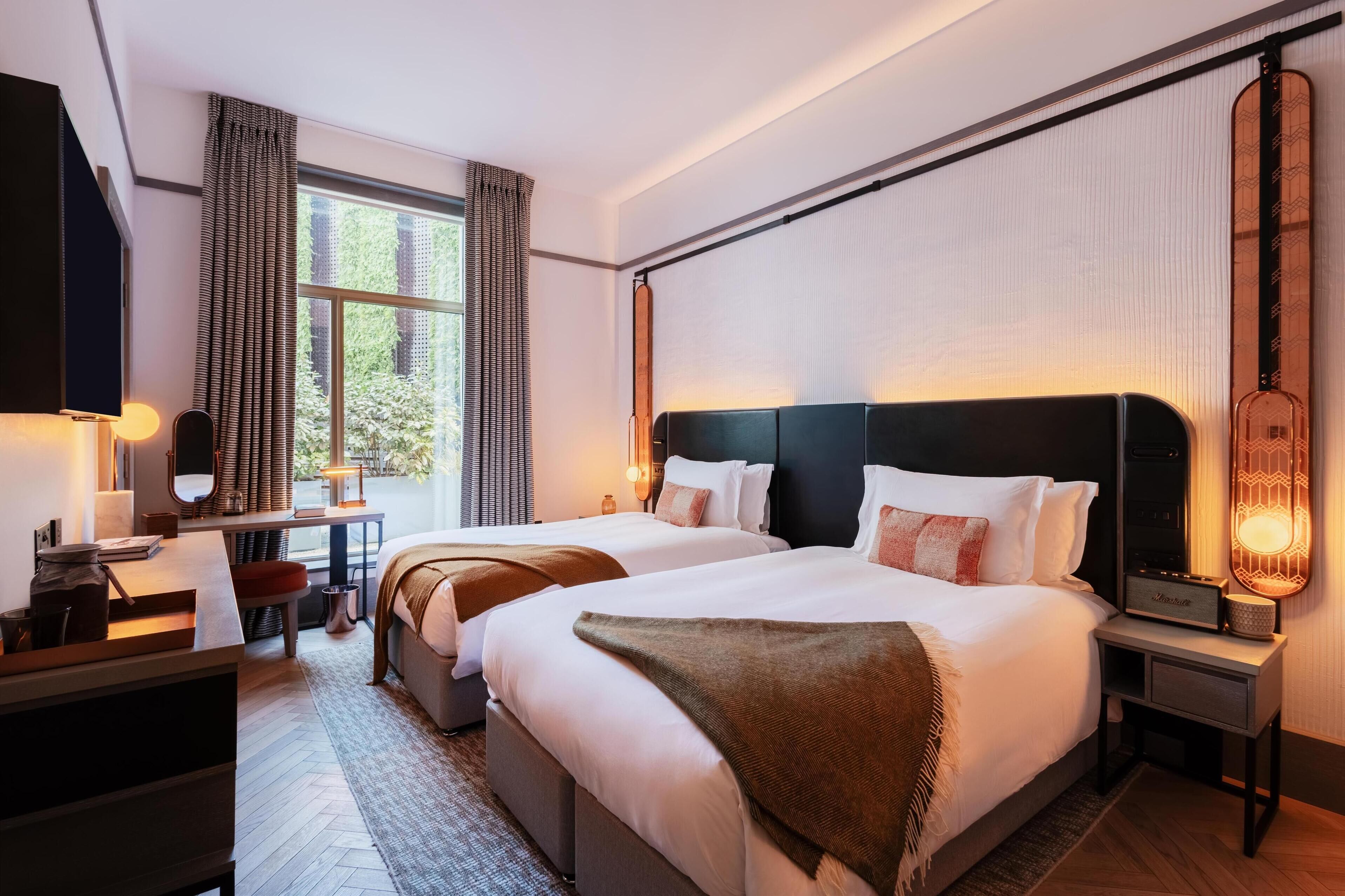 Ett hotellrum med två sängar, ett skrivbord, en stol och ett stort fönster med gardiner.