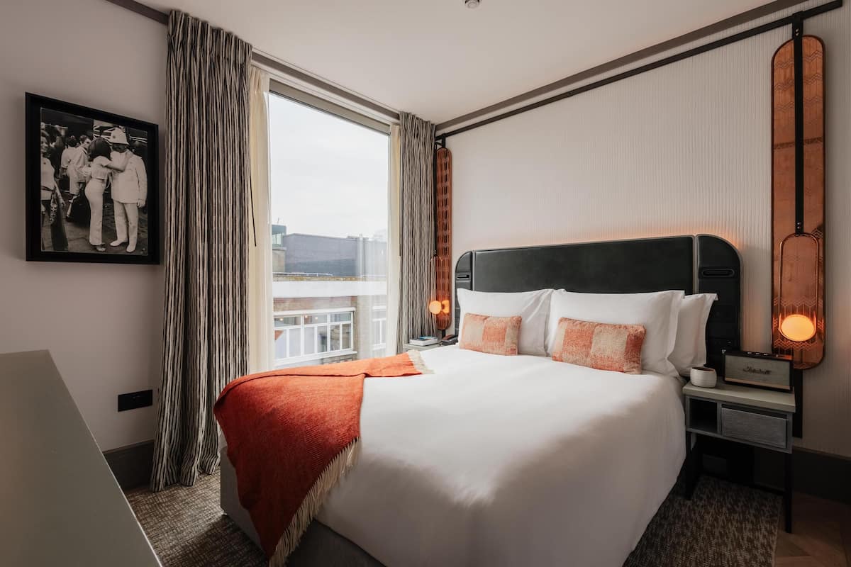 A hotel room with a large bed, a nightstand with a lamp, a framed photo on the wall, and a window with curtains.