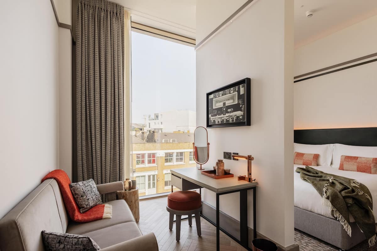 A hotel room with a bed, a desk with a mirror, a window with curtains, a sofa with a red throw, and a city view.