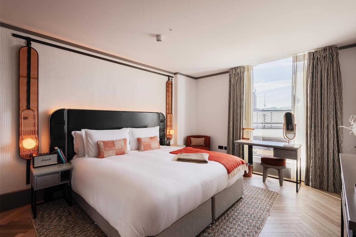 A hotel room with a large bed, a desk with a chair, a window with curtains, and a guitar on the wall.