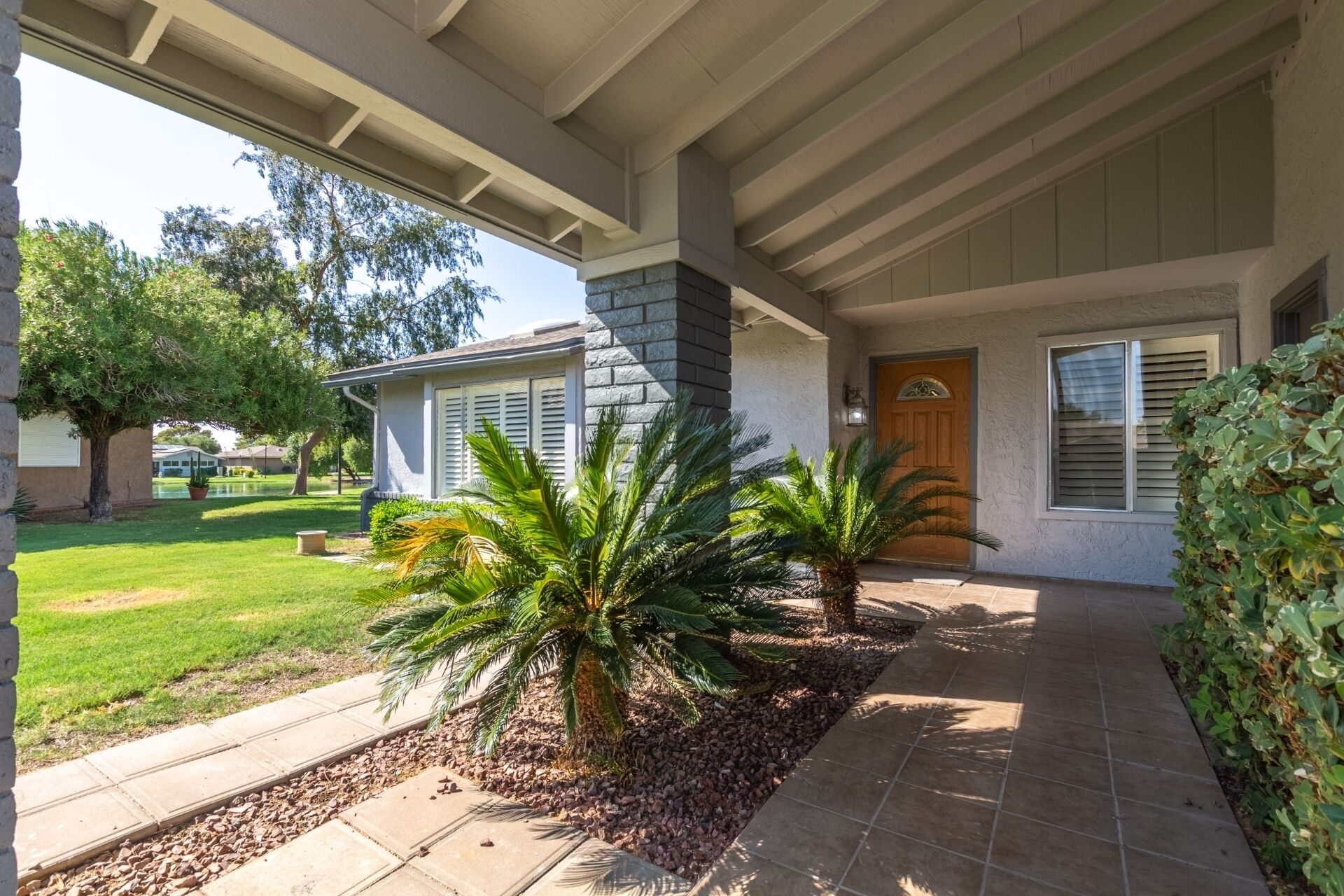 A covered porch with a stone pillar, palm trees, and a pathway leading to a house with a wooden door and shutters.