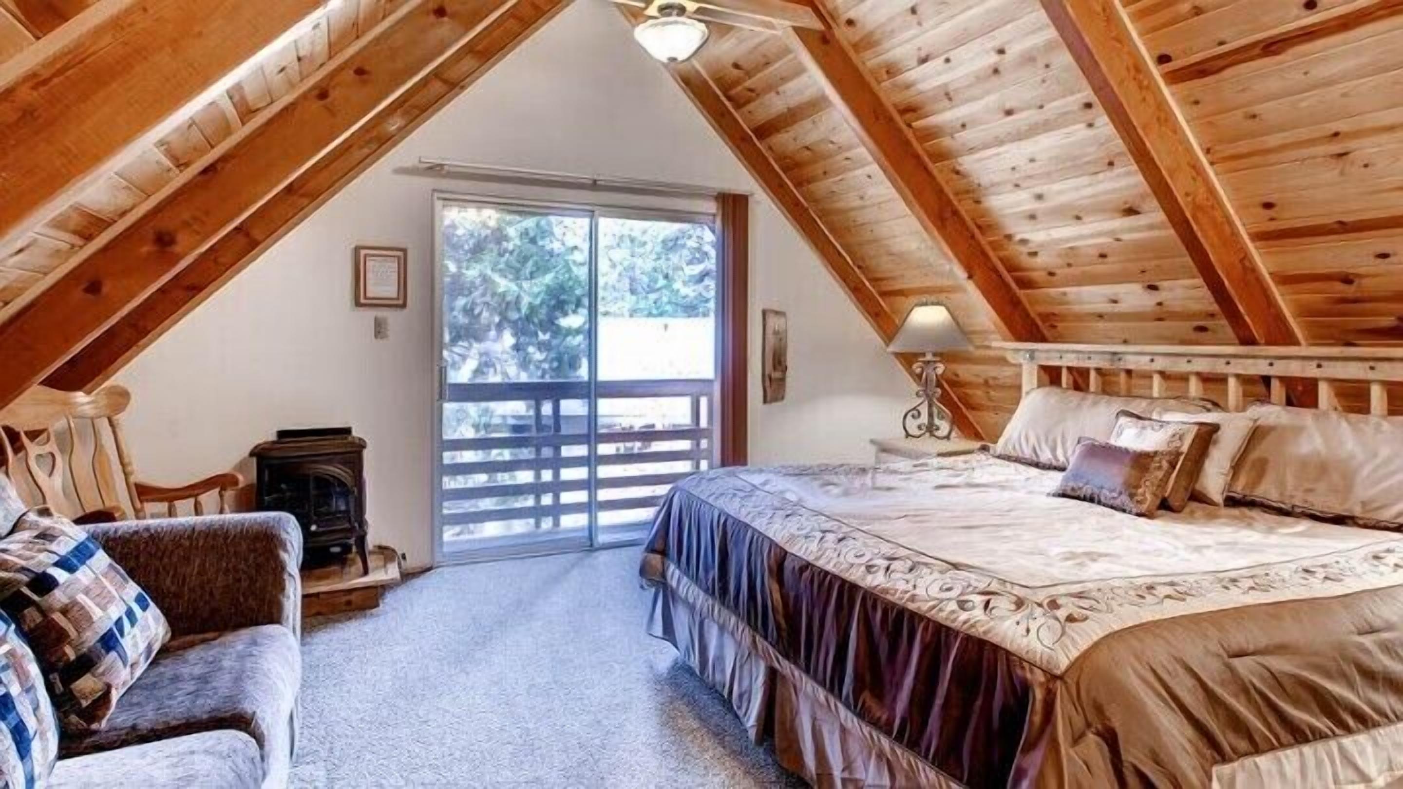 Um quarto de sótão aconchegante com uma cama grande, cabeceira de madeira, uma mesinha com abajur e uma porta de vidro deslizante para uma varanda.