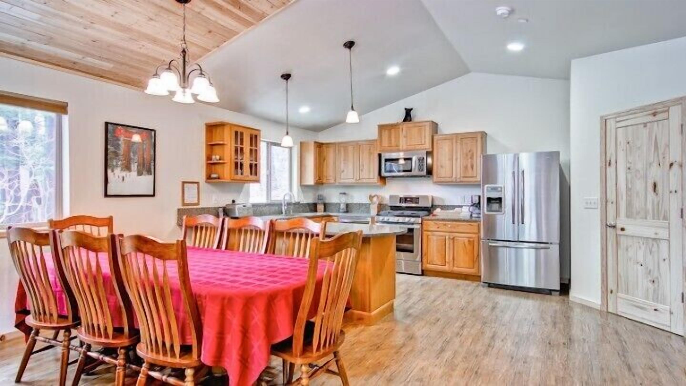 A spacious kitchen with wooden cabinets, a stainless steel refrigerator, and a dining area with a red tablecloth and wooden chairs.