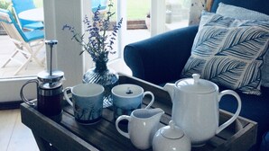 Un salon chaleureux avec une table basse sur laquelle se trouvent une cafetière à piston, une théière et des tasses. On aperçoit un canapé bleu et un coussin à motifs, et de grandes fenêtres offrent une vue sur un jardin.