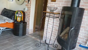 Une pièce avec un mur en briques, un poêle à bois noir, un lit recouvert d’une couverture à motifs, un vieux juke-box et une petite table avec un cadre en métal.