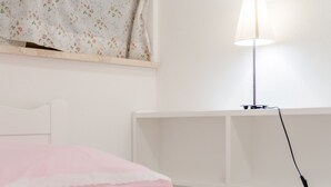 Ein weißes Bett mit einem rosa Kopfkissen, ein weißer Kopfteil, eine weiße Wand, ein weißer Nachttisch und eine weiße Lampe mit weißem Lampenschirm. Ein weißes Kabel ist in die Lampe eingesteckt.