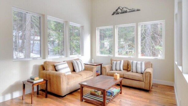 A spacious living room with two sofas, a coffee table, and a side table. Large windows provide a view of trees and a mountain.