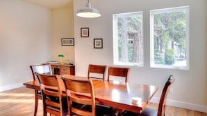 A dining area with a wooden table and chairs, framed pictures on the wall, and two windows with a forest view.