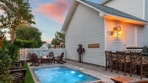 A backyard with a swimming pool, wooden deck, Adirondack chairs, and a white fence.