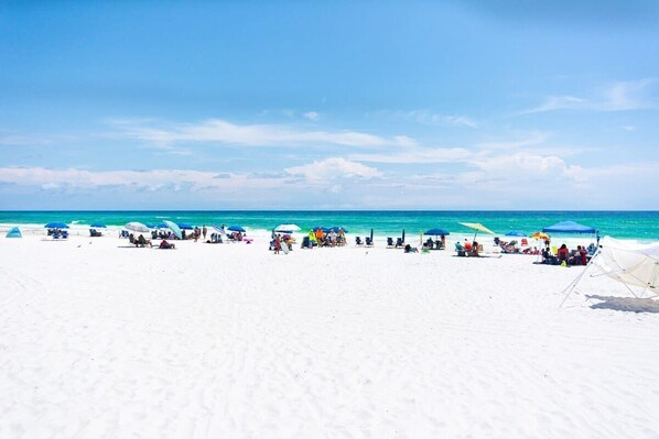 A beach with white sand and clear blue water. There are several umbrellas and people relaxing on the beach.