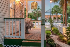 A wooden porch with a white picket fence, a hanging light fixture, and a pathway leading to a white house with a porch.