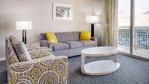 Habitación de hotel con un sofá gris, sillones estampados, una mesa de centro blanca y un ventanal con vistas.