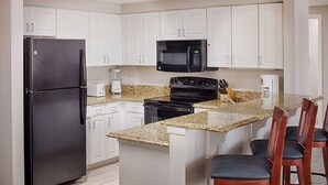 Una cocina con armarios blancos, un refrigerador negro, un microondas, una estufa y una encimera de granito. Hay tres sillas de madera en la isla.