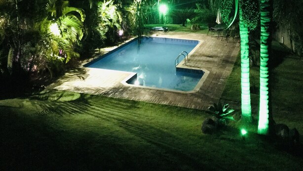 A nighttime outdoor swimming pool surrounded by palm trees and greenery, with a visible pool ladder and a small deck area. - CHÁCARA MARANATA - ATIBAIA | Rentals for Weekends, Holidays and Seasons. (ATIBAIA)