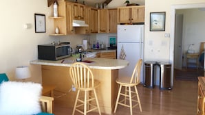 A compact kitchen with wooden cabinets, a microwave, a stove, and a refrigerator. There are two wooden stools and a small table with a lamp. - Mallard Cottage - Pet friendly (Newfield)