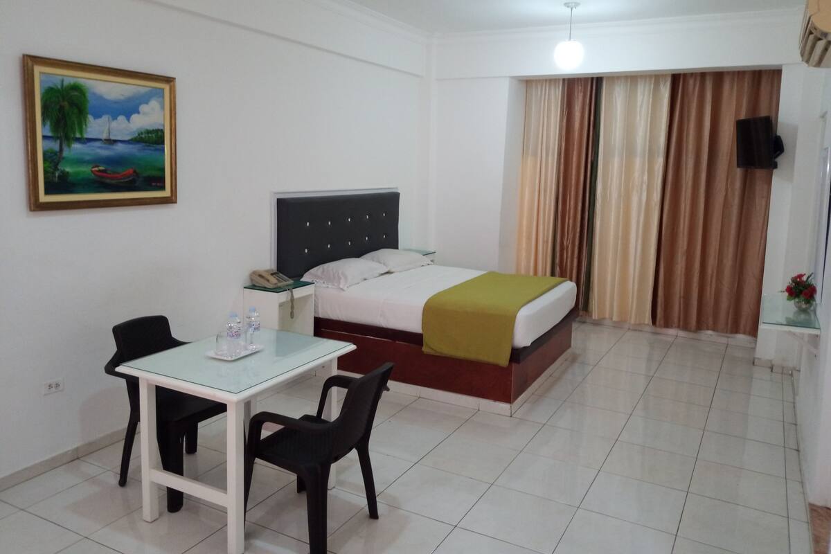 Une chambre d’hôtel avec un lit, un bureau avec des chaises, un tableau au mur et une fenêtre avec des rideaux.