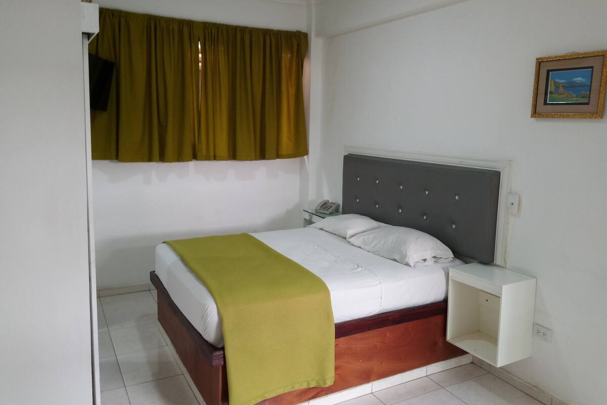 Un lit simple avec une tête de lit rembourrée de couleur grise, un matelas blanc et un plaid vert. Il y a une table de chevet avec un téléphone et une petite étagère. La pièce a des murs blancs, une fenêtre avec des rideaux verts et un tableau encadré au mur.