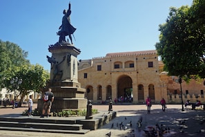 A plaza with a statue, steps, and a building with arches. - Hotel Royal Palace (Santo Domingo)