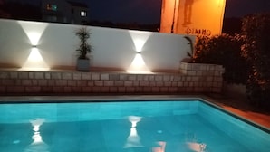 Une piscine bordée de briques et un mur blanc orné d’un motif géométrique. Deux plantes en pot sont disposées sur le mur.