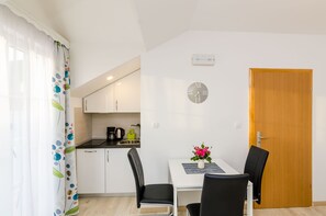 A small, modern kitchen with white cabinets, a black countertop, and a coffee maker. There is a dining table with two chairs and a vase of flowers. A wooden door and a wall clock are also visible.