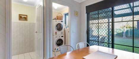 A laundry room with stacked washers and dryers, a table with a placemat, and a view through a window with a metal fence.