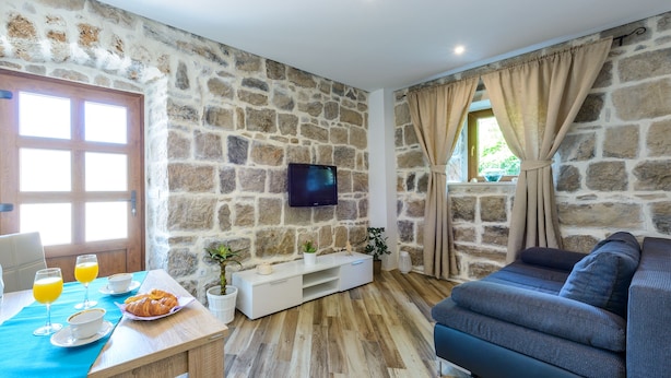 A room with stone walls, a wooden floor, a sofa, a TV, a dining table with breakfast items, and a window with curtains.