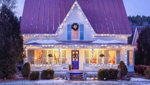 Uma casa de dois andares com telhado vermelho, varanda com luzes de Natal e uma guirlanda na porta da frente.