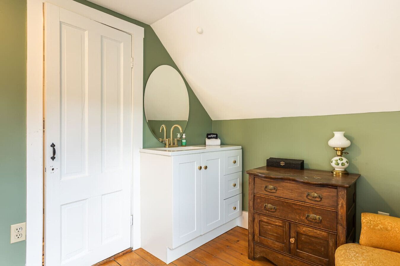 Una habitación con una puerta blanca, un aparador blanco con cajones, un espejo, una mesita con una lámpara y una silla amarilla.