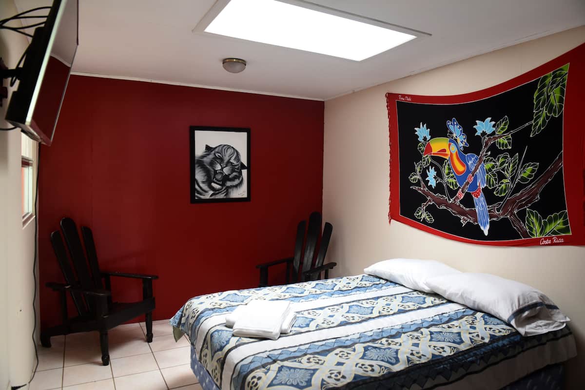Ein Zimmer mit einem Bett, zwei Stühlen, einer Deckenleuchte, einem Wandbild und einem Fenster.