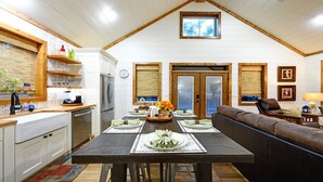 Une salle à manger avec une table en bois dressée pour six personnes, une cuisine avec des meubles blancs et un salon doté d’un canapé en cuir marron.