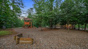Un parque infantil con un módulo de juegos de madera, rodeado de árboles y cubierto de guijarros.