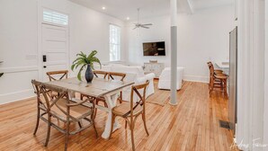 A modern living room with a dining area, white walls, wooden flooring, and a ceiling fan. - 2 King Bedrooms in Historic Barracks at North Beach (Tybee Island)