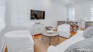 A modern living room with a ceiling fan, a flat-screen TV, a white sofa, a round coffee table, and a dining area with chairs. - 2 King Bedrooms in Historic Barracks at North Beach (Tybee Island)