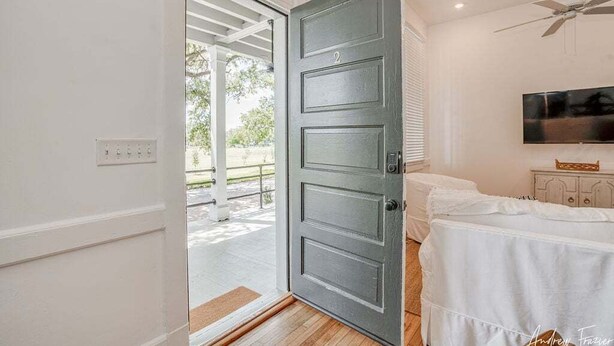 A bedroom with a large dark wooden door, a bed with white bedding, a ceiling fan, and a television on the wall. - 2 King Bedrooms in Historic Barracks at North Beach (Tybee Island)