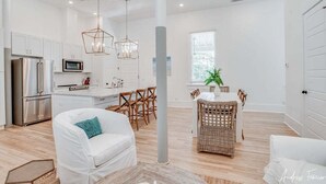 A spacious living area with a white sofa, dining table, and chairs, a kitchen with modern appliances, and a large window providing natural light. - Luxury Tybee Condo with 2 King suites and Pool (Tybee Island)